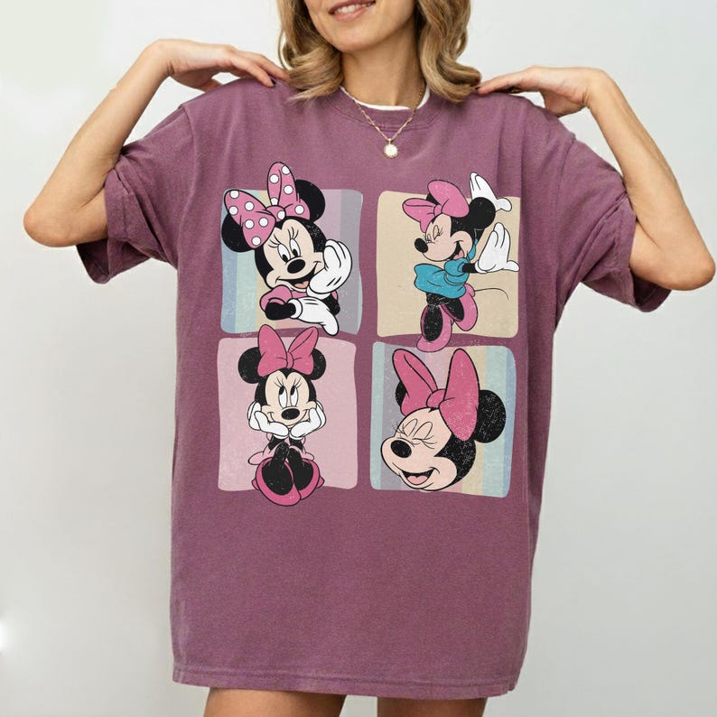 Op de afbeelding: Een mauvekleurig T-shirt met een ontwerp van vier panelen met Minnie Mouse in verschillende poses. Elk paneel heeft een andere achtergrondkleur en Minnie draagt haar kenmerkende roze strik en outfit. Het shirt heeft korte mouwen en een relaxte pasvorm.