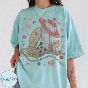 May include: A light blue t-shirt featuring a graphic print of a cowboy boot, a hat, a lasso, and blocks with the letter T and number 13. The text Taylor is on the boot sole, and the text I KNEW IT I KNEW YOU is printed in a circular pattern.