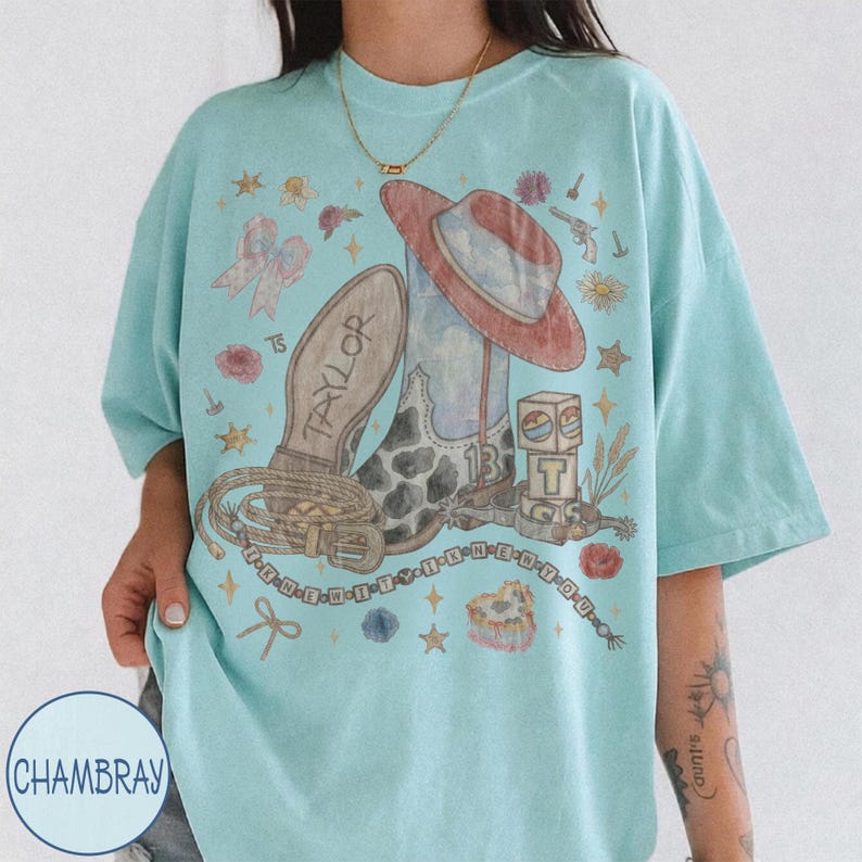 May include: A light blue t-shirt featuring a graphic print of a cowboy boot, a hat, a lasso, and blocks with the letter T and number 13. The text Taylor is on the boot sole, and the text I KNEW IT I KNEW YOU is printed in a circular pattern.