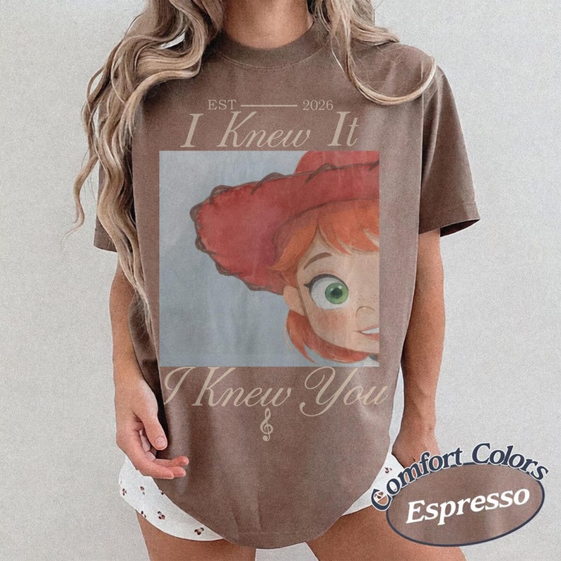 May include: An espresso-colored short-sleeve t-shirt featuring a graphic print of a cartoon character with red hair and a red hat. The text "EST 2026 I Knew It I Knew You" is printed in a light-colored script font above and below the central image.