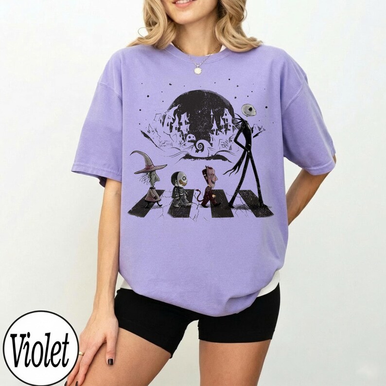 May include: A light violet short-sleeve t-shirt featuring a black graphic print of characters walking across a crosswalk. The design includes a tall skeletal figure, a witch, and two smaller figures against a backdrop of a large moon.