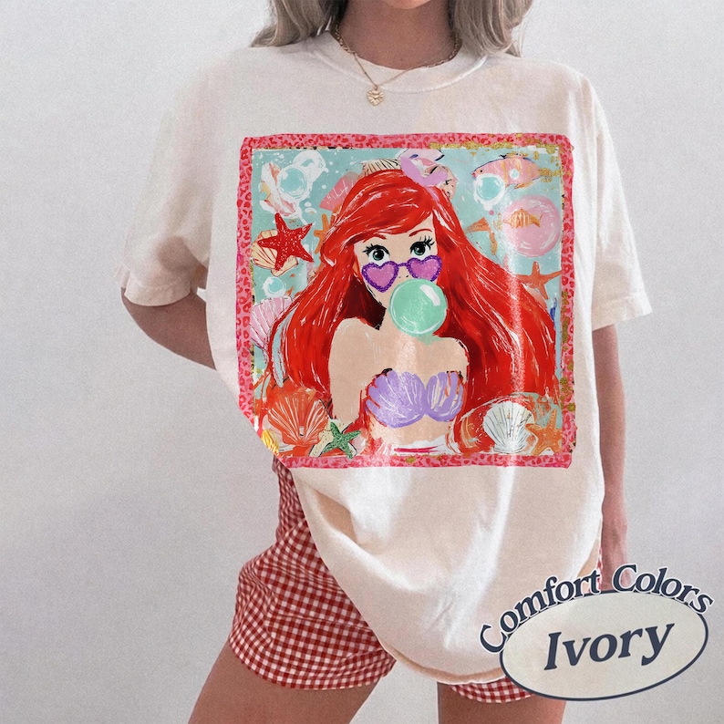 May include: Ivory-colored t-shirt featuring a colorful graphic of a mermaid with red hair, heart-shaped sunglasses, and a bubble. The graphic is surrounded by seashells and starfish. The shirt is labeled "Comfort Colors Ivory".