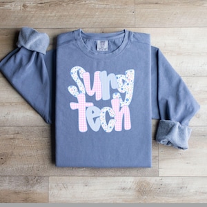 May include: A blue long-sleeve sweatshirt with the words "Surg Tech" in a decorative font. The letters are a mix of floral, gingham, and striped patterns. The sleeves are cuffed.