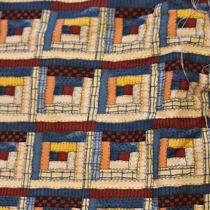 Pode incluir: Um tecido com um padrão repetitivo de blocos de colcha log cabin em tons de azul, amarelo, laranja e vermelho escuro. O design inclui texturas de xadrez e grade. O texto na ourela diz: Y STORE BY DEB STRAIN FOR MODA.