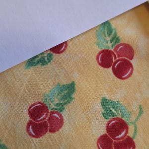 May include: A yellow fabric featuring a repeating pattern of small red berries with green leaves. The berries are arranged in clusters of three, and the yellow background has a subtle, mottled texture throughout the textile design.