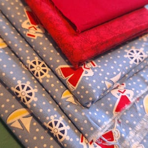 May include: A stack of fabric pieces in shades of red and blue. The blue fabric features a nautical pattern with white polka dots, anchors, and ship wheels. The red fabric is a solid color.