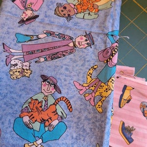 Pode incluir: Pacotes de tecido com ilustrações fantasiosas de figuras com gatos e chitas sobre um fundo azul claro. Um tecido rosa com ilustrações de sapatos também é visível. Os tecidos estão empilhados sobre uma base de corte verde.
