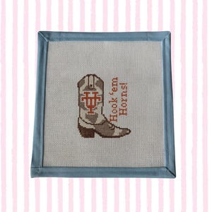 May include: A square needlepoint canvas featuring a white mesh background with a blue fabric border. The center displays a brown and white cowboy boot with a burnt orange logo and the text Hook 'em Horns! stitched in burnt orange thread.