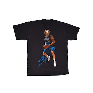 May include: Black t-shirt featuring a basketball player in a blue uniform, holding a basketball. The player's signature is in blue. The jersey number is 41. The shirt is a crew neck.