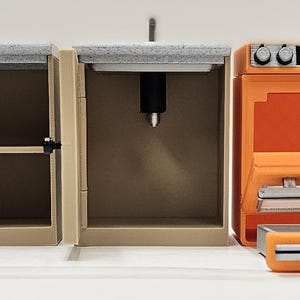 May include: A miniature kitchen set with tan cabinets, a gray countertop, and an orange oven. The oven displays "12:30PM" on its digital clock. The cabinet doors are open, revealing shelves and interior details.