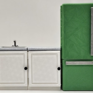 May include: A miniature kitchen set featuring a green refrigerator with silver handles and a white sink cabinet with a gray countertop. The cabinet has two white doors with black knobs. The set is designed for play.