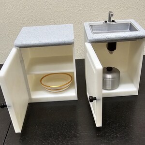 May include: Miniature white kitchen cabinets with open doors. One cabinet has a gray countertop and a gold-rimmed bowl on a shelf. The other cabinet has a sink with a faucet and a silver pot inside. Black knobs and hinges.