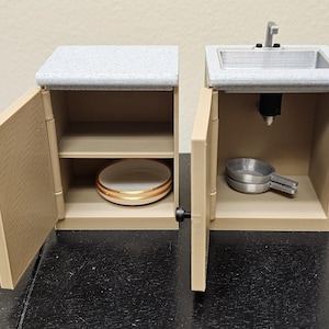 May include: Miniature kitchen set with two cabinets. One cabinet has a sink and faucet, the other has a shelf with a plate. The cabinets are beige with a gray countertop. The plate has a gold rim. The sink is silver.