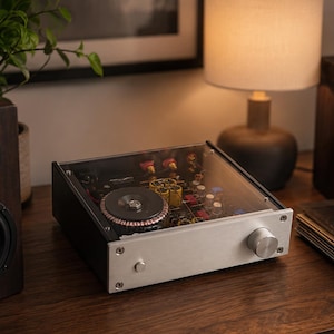 May include: A rectangular audio amplifier featuring a brushed silver metal front panel with a single circular volume knob and a small power button. The top cover is transparent, revealing internal electronic components including a large toroidal transformer.