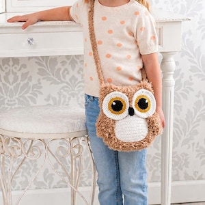 Könnte beinhalten: Eine braun-weiße Umhängetasche in Eulenform mit großen gelb-schwarzen Augen. Die Tasche besteht aus einem weichen, flauschigen Material und hat einen langen Riemen. Die Tasche wird von einer Person getragen, die ein cremefarbenes Polka-Dot-Shirt und eine blaue Jeans trägt.