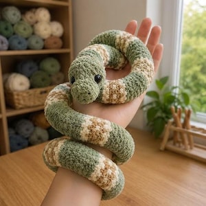 May include: A coiled crochet snake toy featuring alternating bands of sage green and cream yarn. The plush snake has a textured, soft appearance with two small, round, black plastic eyes positioned on its head, creating a realistic look.