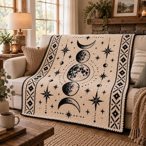 Puede incluir: Una manta de color crema con un diseño estampado en negro de las fases lunares y varias formas de estrellas. La manta tiene un borde decorativo con un patrón geométrico de diamantes repetido a lo largo de los bordes verticales.