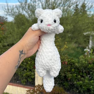 Puede incluir: Un peluche de hurón blanco hecho a mano con ganchillo. El hurón tiene un cuerpo esponjoso, ojos negros pequeños, una nariz rosa y brazos y piernas diminutos. El juguete se sostiene sobre un fondo de follaje verde y un cielo nublado.