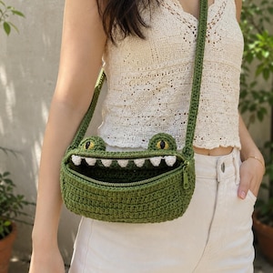 May include: A green crocheted crossbody bag shaped like a crocodile head with a wide open mouth, white triangular teeth, and yellow eyes. The bag features a zipper closure along the top edge of the mouth and a long, matching green crocheted strap.