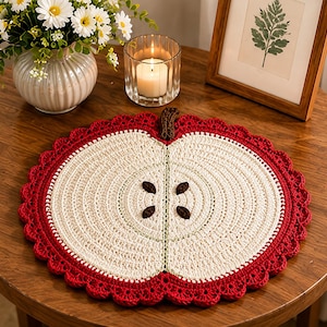 May include: A round, crocheted placemat shaped like a sliced apple. The center is cream-colored with dark brown seed details, surrounded by a vibrant red border with a scalloped edge. A small brown crocheted stem is attached to the top edge.