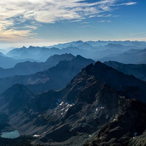 Op de afbeelding: Een landschap van bergketens onder een blauwe lucht met verspreide wolken. De bergen zijn in verschillende blauwtinten, wat een gevoel van diepte en afstand creëert. Een klein meer is zichtbaar in de vallei.