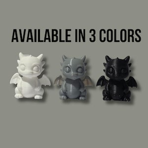 May include: Three small dragon figurines in white, gray, and black. Each creature features large eyes, pointed ears, and small wings. The text "AVAILABLE IN 3 COLORS" is printed in black above the figures, which are arranged in a horizontal row.