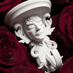 Puede incluir: Un aplique de pared blanco y ornamentado con un rostro esculpido y detalles florales, sobre un lecho de rosas rojas oscuras. El aplique presenta una parte superior en forma de sombrero y detalles intrincados.