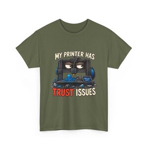 Pode incluir: Uma t-shirt verde-oliva com um gráfico de uma impressora 3D com olhos zangados e um pequeno polvo azul na base. O texto diz "MY PRINTER HAS TRUST ISSUES" com "Are you sure?" no ecrã da impressora em branco e vermelho.