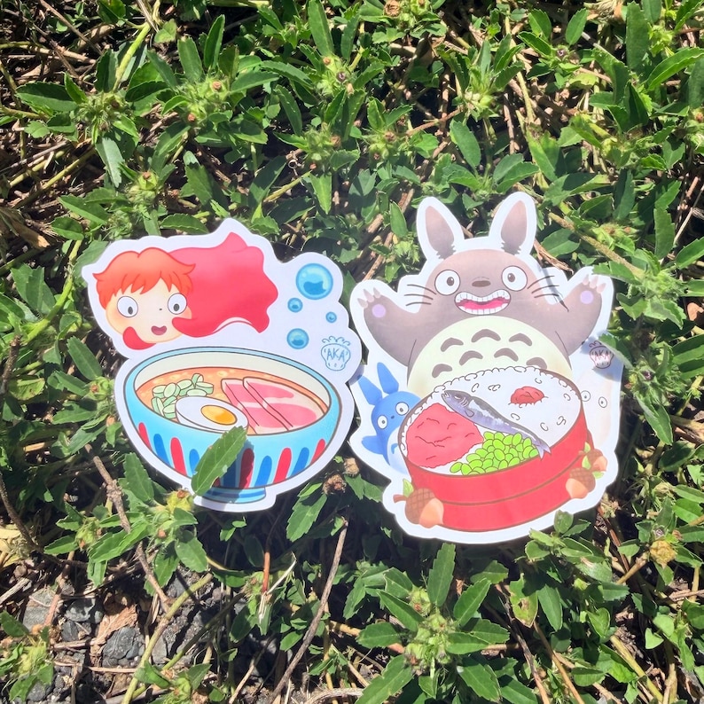 Puede incluir: Dos pegatinas coloridas con ilustraciones de estilo anime. Una muestra a una chica con cabello rojo y un cuenco de ramen. La otra representa una criatura grande, gris, parecida a un conejo, con una caja bento y una peque&ntilde;a criatura azul.