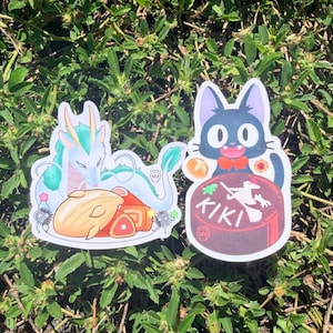 Puede incluir: Dos pegatinas con ilustraciones de estilo anime. Una representa un drag&oacute;n turquesa con un plato de comida. La otra muestra un gato negro con pajarita roja, sentado sobre una tarta con la palabra "KIKI".