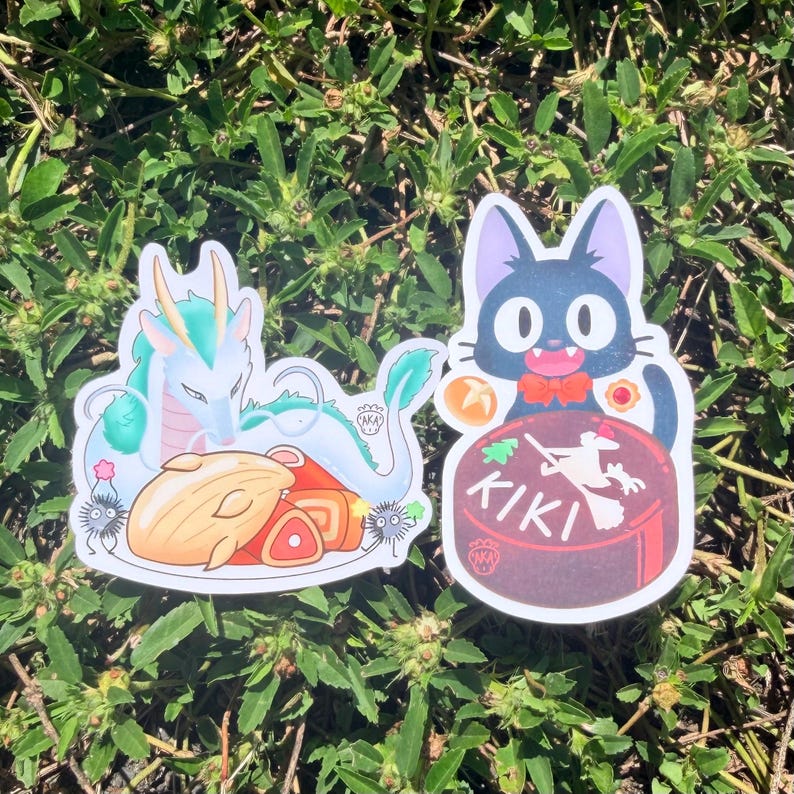 Puede incluir: Dos pegatinas con ilustraciones de estilo anime. Una representa un drag&oacute;n turquesa con un plato de comida. La otra muestra un gato negro con pajarita roja, sentado sobre una tarta con la palabra "KIKI".
