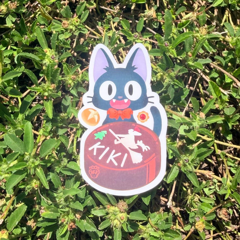 Puede incluir: Una pegatina con un gato negro con ojos grandes y una pajarita roja. El gato sostiene un pastel marr&oacute;n con la palabra "KIKI" y una bruja en una escoba. La pegatina est&aacute; sobre un lecho de plantas verdes.