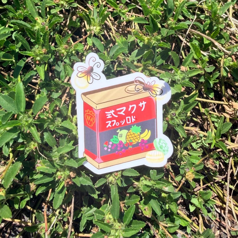 Puede incluir: Una pegatina con un dise&ntilde;o de caja rectangular con texto japon&eacute;s y una ilustraci&oacute;n de frutas. La caja es roja y dorada, con un borde negro y un colorido arreglo de frutas que incluye uvas, pi&ntilde;a y pl&aacute;tanos. La pegatina est&aacute; sobre follaje verde.