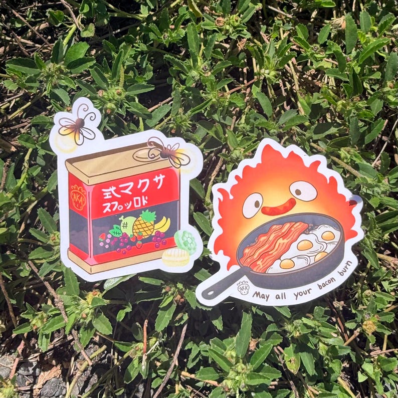 Puede incluir: Dos pegatinas sobre un lecho de follaje verde. Una es una caja roja y dorada con texto japon&eacute;s e ilustraciones de frutas. La otra es una caricatura de un fuego con una sart&eacute;n de tocino y huevos, con el texto "May all your bacon burn."
