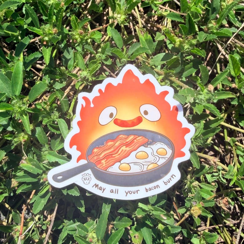 Puede incluir: Una pegatina con una llama de dibujos animados con una cara sonriente sobre una sart&eacute;n con tocino y huevos. El texto "May all your bacon burn" est&aacute; escrito debajo. La pegatina est&aacute; sobre hierba verde.
