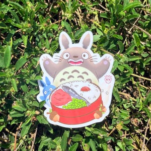 Puede incluir: Una pegatina troquelada con un personaje de dibujos animados de Totoro con una caja bento. La pegatina tiene un Totoro marr&oacute;n con una cara sonriente, un personaje azul y una caja bento roja y blanca con comida.