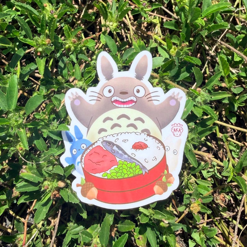 Puede incluir: Una pegatina troquelada con un personaje de dibujos animados de Totoro con una caja bento. La pegatina tiene un Totoro marr&oacute;n con una cara sonriente, un personaje azul y una caja bento roja y blanca con comida.
