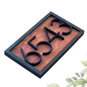 Puede incluir: Letrero de dirección rectangular con los números 6543 en negro, montados sobre un fondo de madera teñida. Los números están en relieve y tienen una fuente moderna. El letrero está enmarcado en un borde gris oscuro o negro.