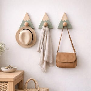 Puede incluir: Tres percheros de pared con forma de montaña con un sombrero, una bufanda y un bolso colgados. Los percheros están pintados en colores neutros y tienen una bola de madera en la base. El sombrero es marrón claro, la bufanda es beige claro y el bolso es marrón.