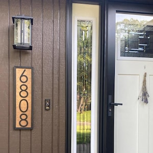 May include: A brown wooden house number sign with the number 6808 in a black font. A black outdoor light fixture is mounted above the sign. The door is white with a black frame and a glass panel.