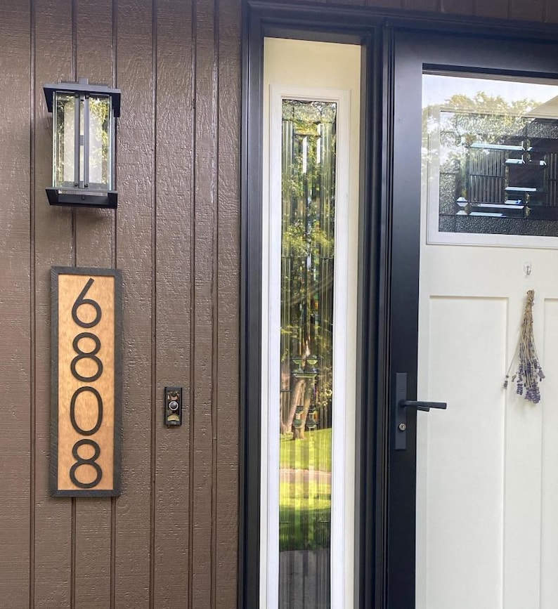 May include: A brown wooden house number sign with the number 6808 in a black font. A black outdoor light fixture is mounted above the sign. The door is white with a black frame and a glass panel.