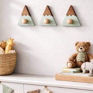 May include: Three wooden mountain-shaped wall hooks with a light green base, brown peak, and a natural wood knob. A woven basket with stuffed animals, a teddy bear, and a garland of wooden beads decorate the room.