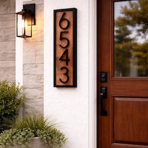 May include: A wooden address sign with the numbers 6543 in a black frame. The sign is mounted on a white wall next to a dark wooden door. A black lantern and a potted plant are also visible.
