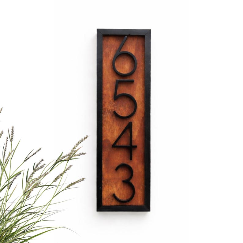 May include: Vertical wooden address sign with the numbers 6543 in black against a stained wood background. The numbers are in a modern font and are framed by a black border. The sign is mounted on a white wall.