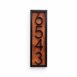 May include: Vertical wooden address sign with the numbers 6, 5, 4, and 3 in black. The numbers are stacked vertically on a brown wooden background, framed by a black border. The sign is rectangular and designed for outdoor use.
