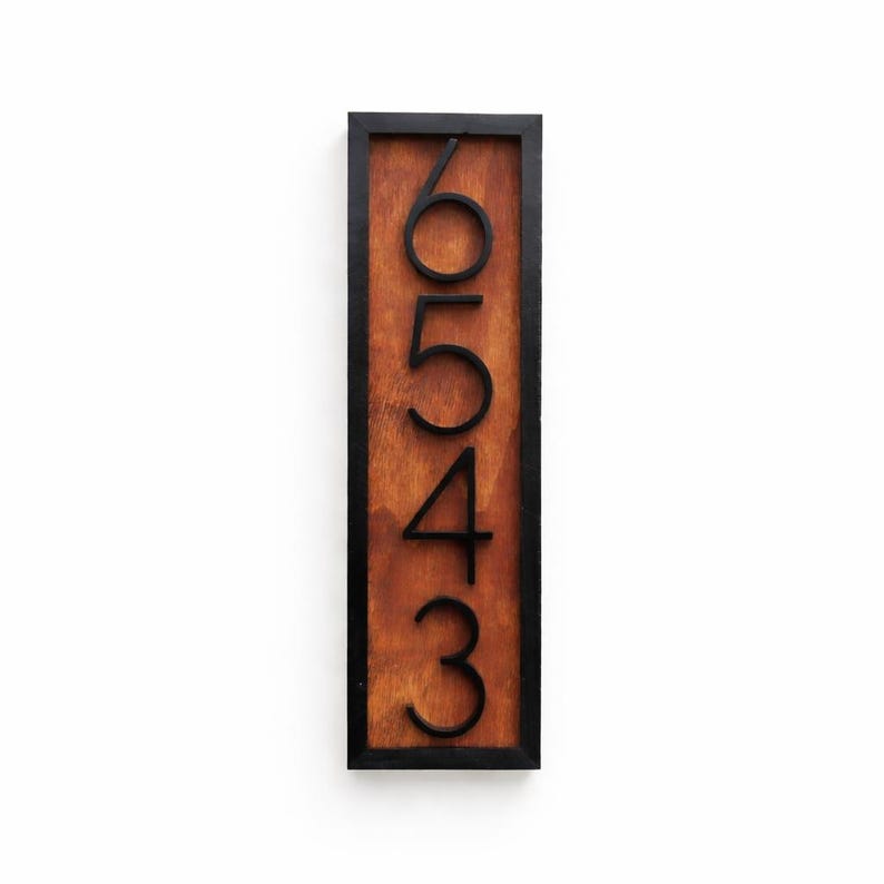 May include: Vertical wooden address sign with the numbers 6, 5, 4, and 3 in black. The numbers are stacked vertically on a brown wooden background, framed by a black border. The sign is rectangular and designed for outdoor use.