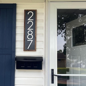 May include: A rectangular wooden address sign with the numbers "2787" in white. The sign is mounted on a white clapboard wall next to a black mailbox and a white door with a glass panel. A dark blue shutter is on the left.