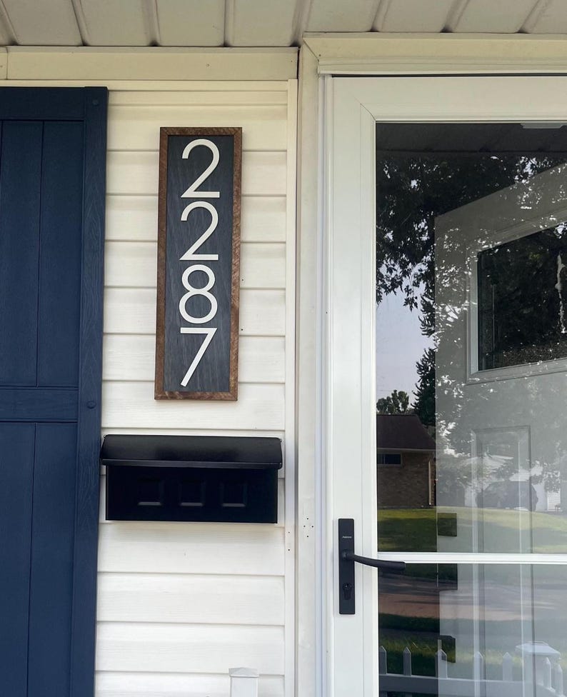 May include: A rectangular wooden address sign with the numbers "2787" in white. The sign is mounted on a white clapboard wall next to a black mailbox and a white door with a glass panel. A dark blue shutter is on the left.