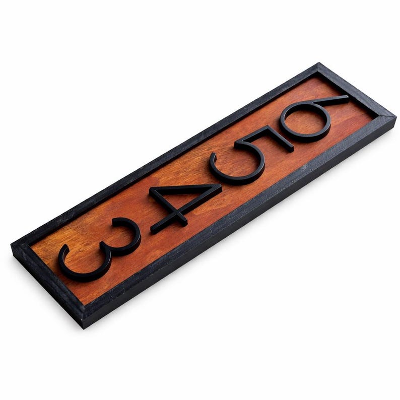 May include: A rectangular address sign with the numbers 3456 in black, mounted on a stained wooden board. The board is framed in black, creating a contrast with the warm wood tones. The numbers are modern and three-dimensional.