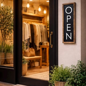 Puede incluir: Una fachada de tienda con una puerta negra y un letrero que dice "OPEN" en letras blancas. La puerta refleja el interior de una tienda de ropa con percheros. Cestas de mimbre con plantas están frente a la puerta.
